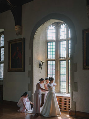 Dramatic candid photography of bridesmaids assisting the bride in a wood-paneled room with a large arched window.