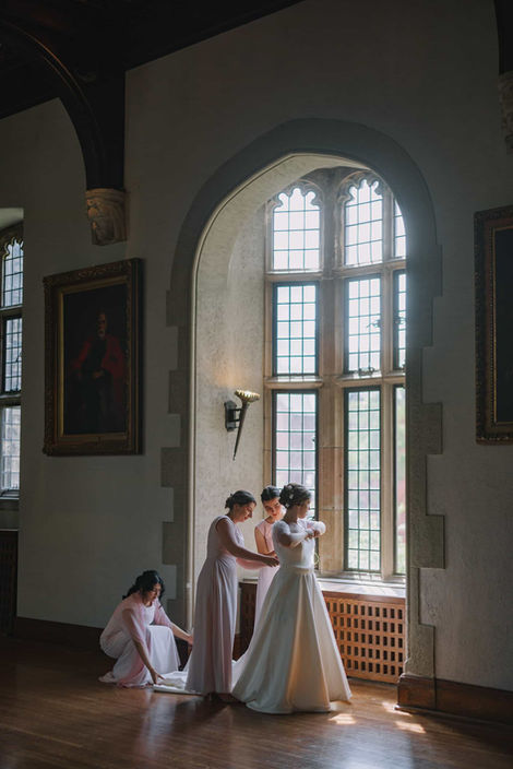 Dramatic candid photography of bridesmaids assisting the bride in a wood-paneled room with a large arched window.