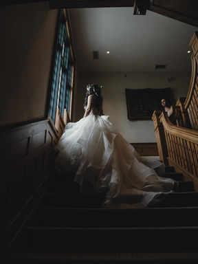 A dramatic, moody bridal portrait from behind a bride in a full ballgown looking up a dark staircase toward window light.