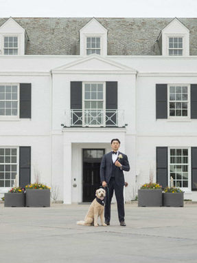 A groom in a classic black tuxedo posing with his dog in front of a white luxury estate.