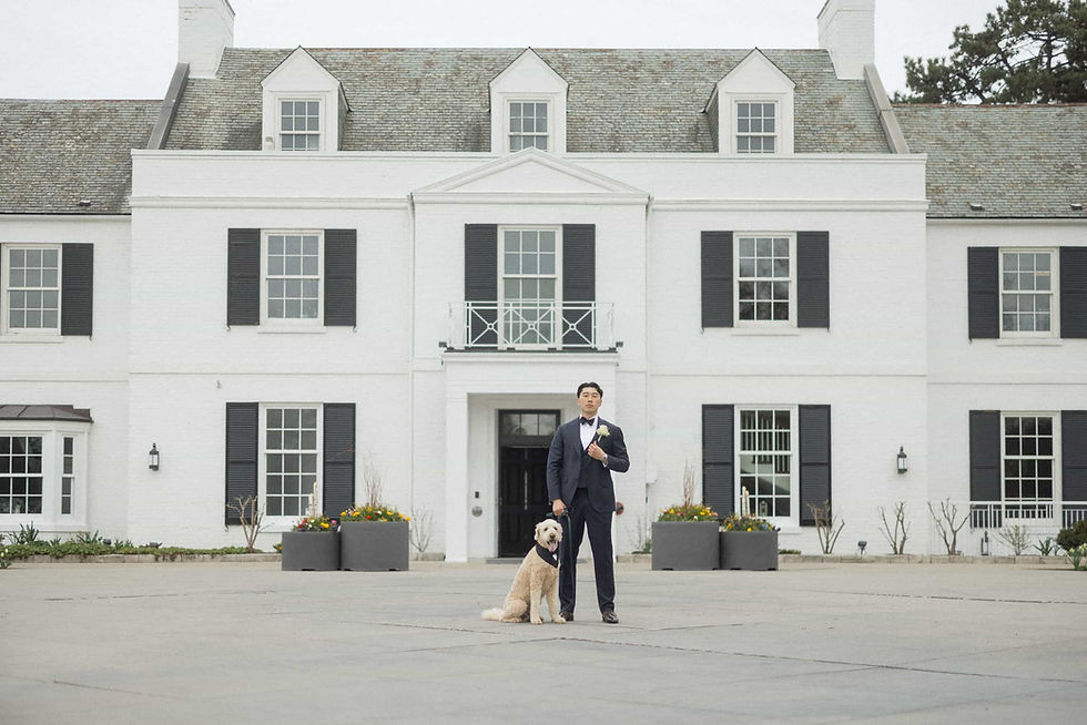 A man in a tuxedo stands with a dog in front of a large white house with black shutters. The setting is elegant and formal.