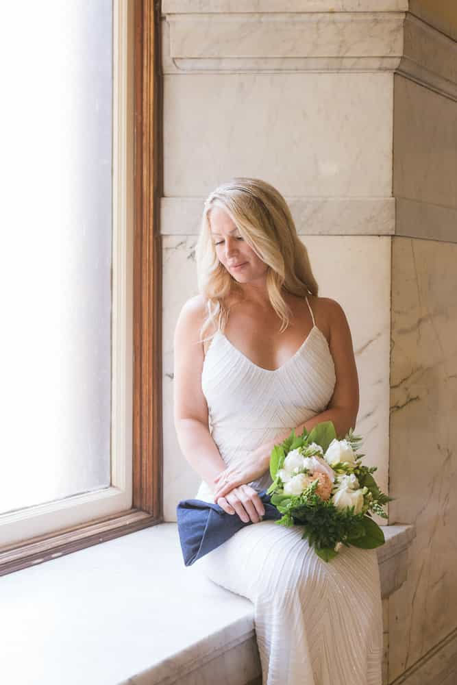 A woman in a white dress holding flowers sits by a large window in a marble setting, looking down thoughtfully. Soft lighting.