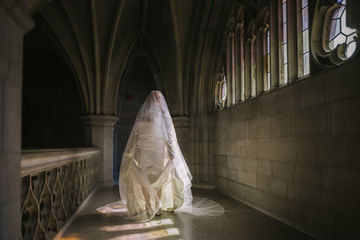 An artistic portrait of a bride fully draped in a long flowing veil, standing in the dramatic shadows and light rays of a stone gothic hallway.


