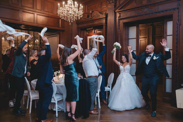 A bride, groom, and their bridal party cheering with their hands in the air during a lively grand entrance in a classic wood-paneled room.