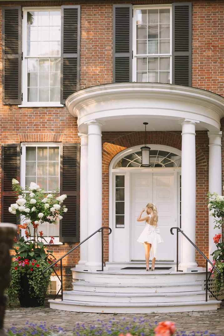 A woman in a white dress poses happily at the top of the steps in front of the white door of a classic Toronto townhouse.