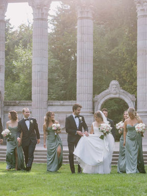 A bright, sun-flared wide shot of the wedding party walking in front of the sweeping stone pillars at The Guild Inn Estate.