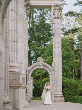 A distant storytelling wedding photograph of a bride walking gracefully beneath a soaring, historic stone arch monument.