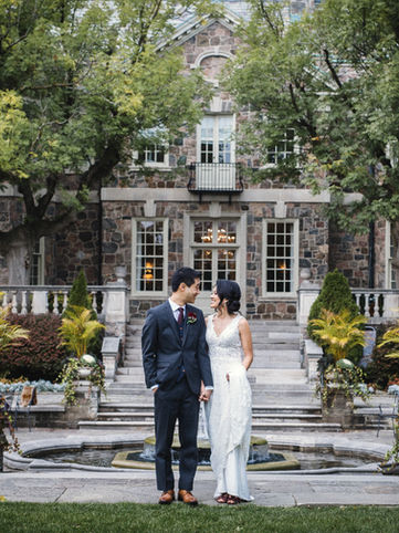 A wide portrait of a newlywed couple posing on a stone terrace courtyard in front of the grand stone steps and formal gardens of the manor.