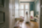 Bride in white gown stands in an elegant teal dressing room with tall windows, mirrors, and pink chairs.