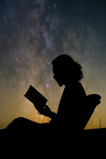 Silhouette of a person reading a book against a starry night sky transitioning from blue to gold.