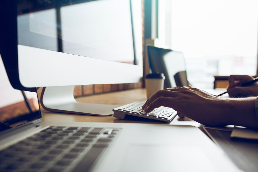 Close-up of hands typing on a laptop keyboard at a desk, computer monitor in the background