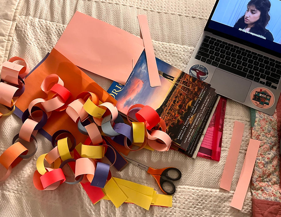 on a white bedspread, there is a laptop and a pile of multi coloured construction paper. There is also a pair of scissors and a multi coloured paper chain