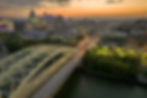 Aerial view of a Cincinnati at sunset with a bridge over a river. Skyscrapers and highways are lit, creating a vibrant urban scene.