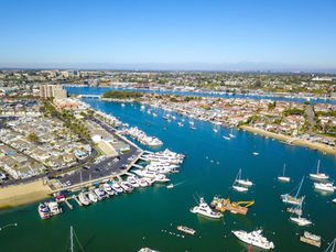 Aerial view of a Newport Beach marina with numerous white boats docked and some sailing on blue water. Residential area with palm trees in the background.