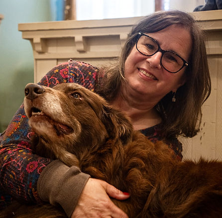 Mindy sits holding her brown dog, both appearing calm and relaxed in a comfortable, home-like setting.