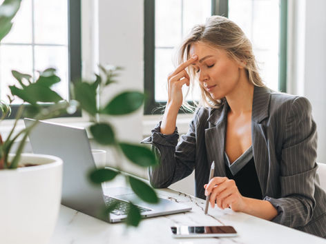 Femeie în costum office, cu semne vizibile de stres, stând la birou în fața laptopului. Imagine asociată cu epuizarea profesională, anxietatea la locul de muncă și dezechilibrul între viața personală și cea profesională. Conținut educativ susținut de Kinetovital, București – Sector 4