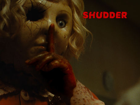 Creepy doll with cracked skin, one eye missing, and a bloodied finger on its lips for silence, set against a dim background. Eerie mood. Shudder logo.