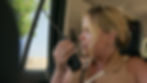 Woman in a beige top holds a walkie-talkie in a car, looking out the window with a concerned expression. Sunglasses are on her head.