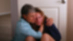 Two people sit on the floor in a comforting embrace. The older person wears a blue patterned shirt, while the younger one appears to be crying.