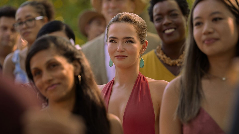 Smiling woman in a pink halter dress and turquoise earrings stands among a blurred crowd outdoors in warm sunlight.