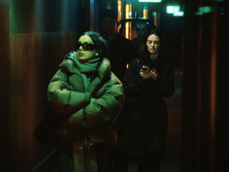 Women walk down a dimly lit hallway. One wears a green puffer jacket and sunglasses, the other checks her phone. Moody atmosphere.
