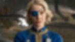 Blonde woman with blue eye patch and uniform stands confidently in industrial setting. Blue and gold accents visible, focused expression.