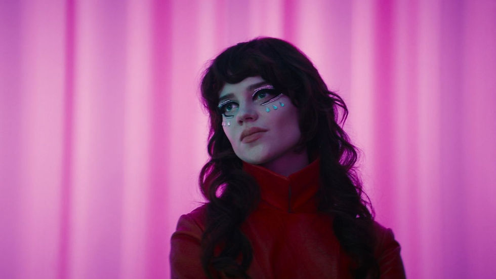 Pensive young woman with glitter tears and dark wavy hair, wearing a red high-collar top against pink curtain backdrop.