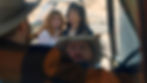 Two women in casual clothes look concerned at a man in a hat inside a car. Background shows a desert road, suggesting a tense desert situation.