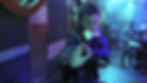 Man in a neon-lit club blocks a blow with a card in his mouth, one hand raised, as blurred people watch in the background.
