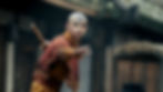 Bald boy in orange robes with a forehead tattoo reaches forward in a martial arts stance, a staff on his back.
