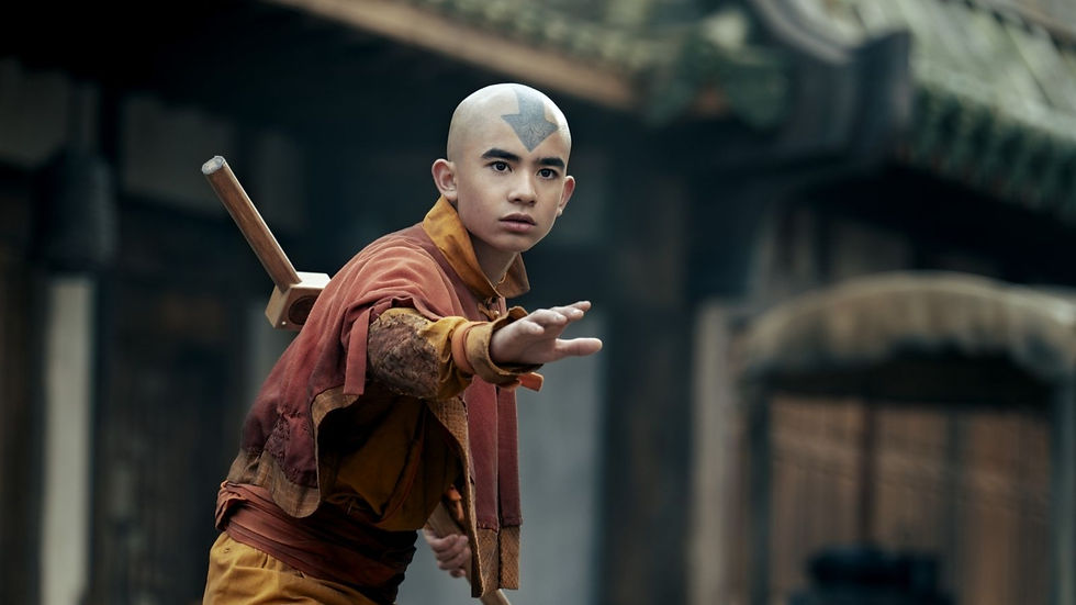 Bald boy in orange robes with a forehead tattoo reaches forward in a martial arts stance, a staff on his back.
