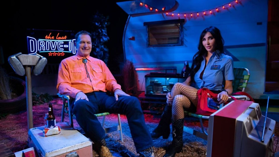 Two people sit outside a retro camper with "The Last Drive-In" sign. One wears orange, the other blue. Red lights create a cozy mood.