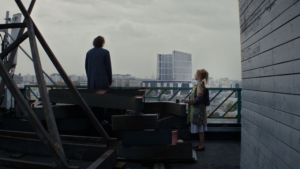 Two people stand on a rooftop with industrial structures, overlooking a city skyline under a cloudy sky. One faces away, the other holds a cup.