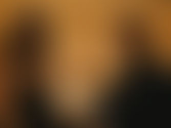 Two people stand at a bar looking concerned. Warm lighting and hanging bulbs create a tense mood. A candle and drink are on the counter.