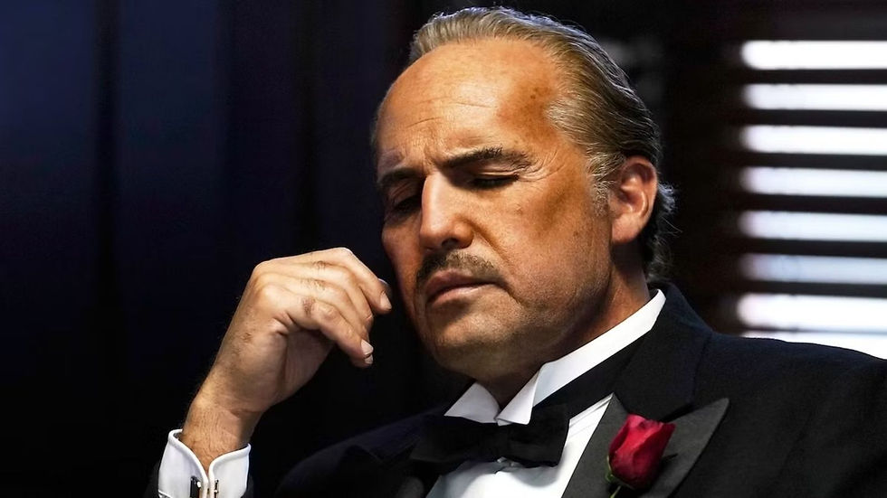 Man in a tuxedo with a red rose, deep in thought, rests hand on face. Dark background with blinds, moody ambiance.