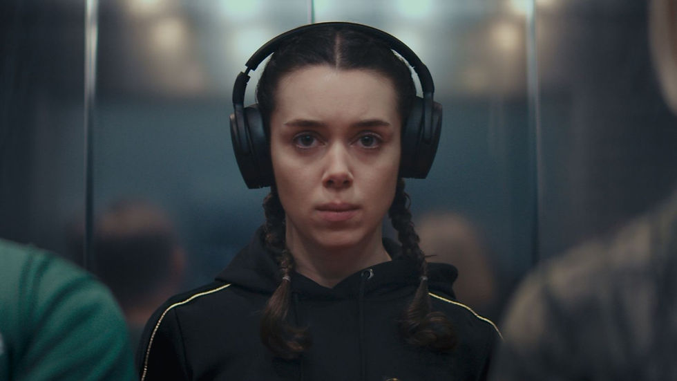 Person with braided hair wearing black headphones and a hoodie, looks focused in an elevator. Blurred figures and metallic walls surround.