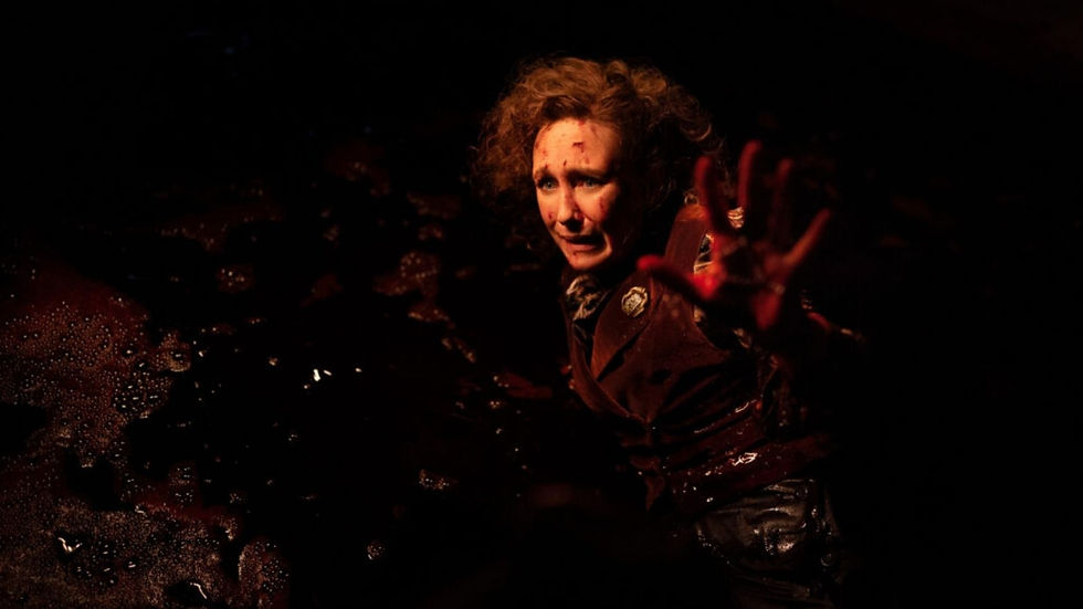 A person with curly hair, covered in dark liquid, reaches out with a red-stained hand in a dimly lit, tense setting.