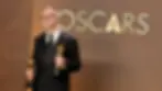 Man in a tuxedo holding two Oscars, smiling in front of a brown backdrop with "OSCARS" text, projecting joy and accomplishment.