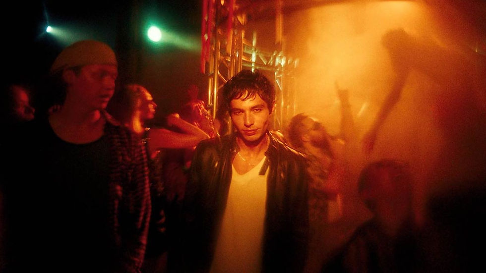 Young man in a leather jacket stands in orange club lights amid a blurry dancing crowd, with a moody, intense vibe.