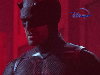 A hero in a black suit stands in a dimly lit red room with crates. The atmosphere is intense and mysterious.