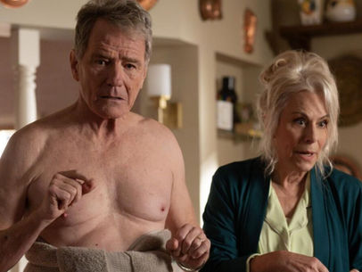 Older man with a towel looks surprised in a cozy kitchen. Woman beside him appears concerned. Warm tones and cookware adorn walls.