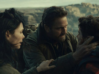 A man holds a teenager's face while a woman watches, all in emotional focus. They're outdoors with cliffs in the distant background.