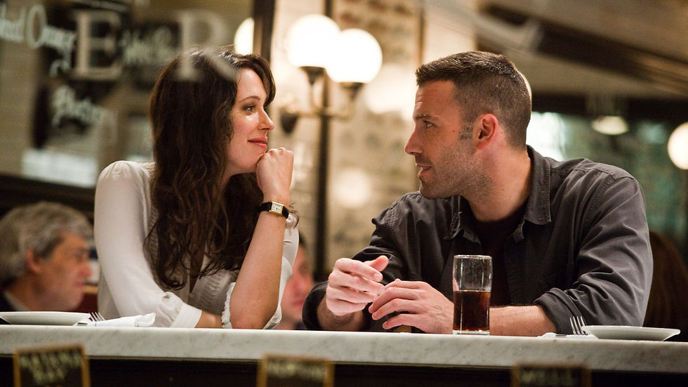 Smiling man and woman chat across a restaurant bar, with a glass of dark drink on the table under warm lights.
