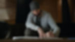 Man wearing a dark cap and gray hoodie writes in a notebook at a wooden table. Dimly lit, cozy room with black and wood background.