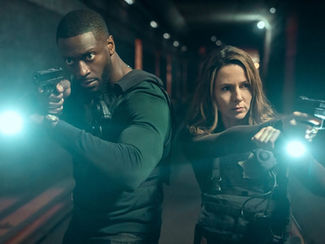 Two people in tactical gear aiming guns with flashlights on, in a dimly lit hallway. They look focused and serious, with blue light accents.