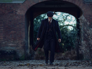Man in dark coat and hat walks under brick archway. Background has trees and dry plants. Mood is serious and mysterious.