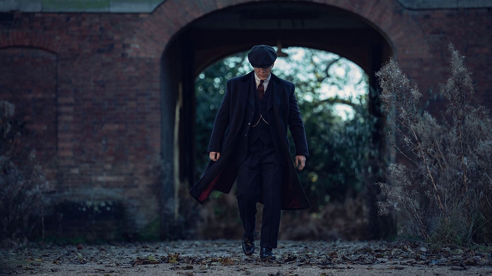 Man in dark coat and hat walks under brick archway. Background has trees and dry plants. Mood is serious and mysterious.