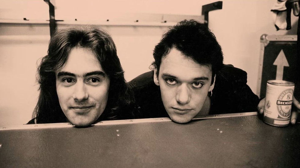 Two men smiling and peering over a table, one holding a Beck's beer can. Behind, a case with an arrow. Black and white photo.