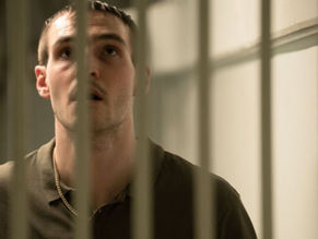 A man in a green shirt looks up behind prison bars, casting a thoughtful expression in a dimly lit cell with a pale background.