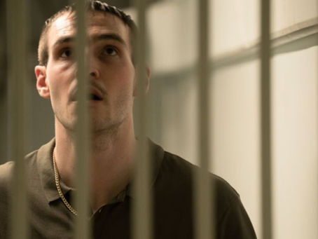 A man in a green shirt looks up behind prison bars, casting a thoughtful expression in a dimly lit cell with a pale background.